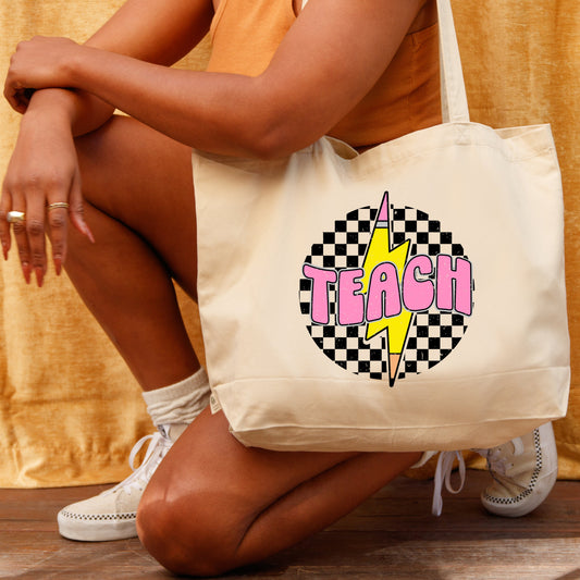 Person holding a tote bag with 'TEACH' design with yellow pencil as a lightning bolt on checkered background against a yellow curtain backdrop