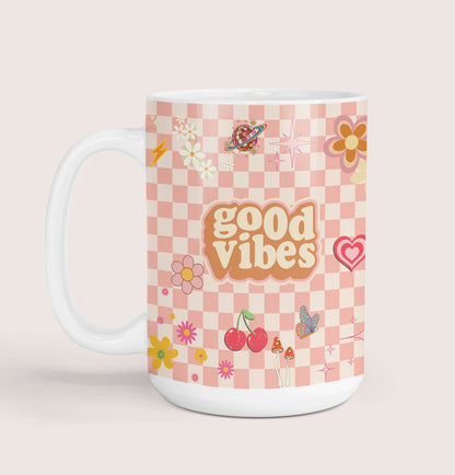 A white ceramic mug with a pink checkered pattern and various colorful illustrations, such as flowers, cherries, mushrooms, butterflies and the text 'good vibes' in the center.